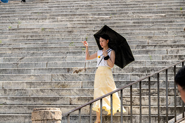 Rom â Lazio â Italy â 08-21-2025: Close view of a woman with a black umbrella, white Shirt and yellow skirt pointing at something, descending on wide marble steps forming a geometric backdrop.