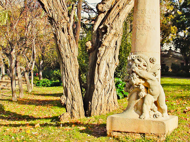 Stone cherub statue decorating a monument in a public park, with tree trunks and fallen autumn leaves on green grass