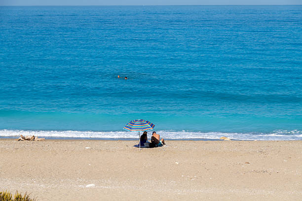 Nydri - Ionian Islands - Greece - 08-09-2025: Two people sit beneath a striped umbrella on a sandy beach, facing the calm blue sea, while two swimmers are visible in the distance.