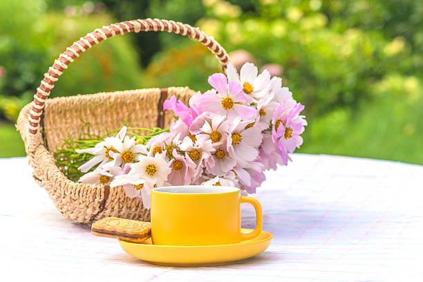 Yellow cup of tea, bouquet of cosmos flowers in a wicker basket on a table with white tablecloth in a blooming garden