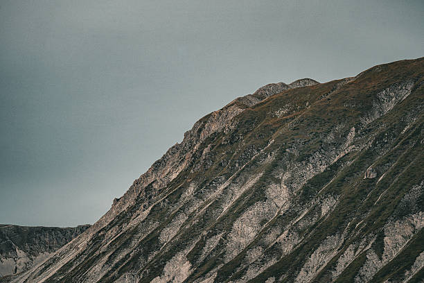 A close-up, dramatic shot focusing on the steep side of the Gran Sasso mountain, showing distinct diagonal rock formations and geological strata interspersed with patches of green vegetation, rising sharply towards the upper ridge against a muted, overcast sky, highlighting the rugged terrain and unique geology of the Apennines