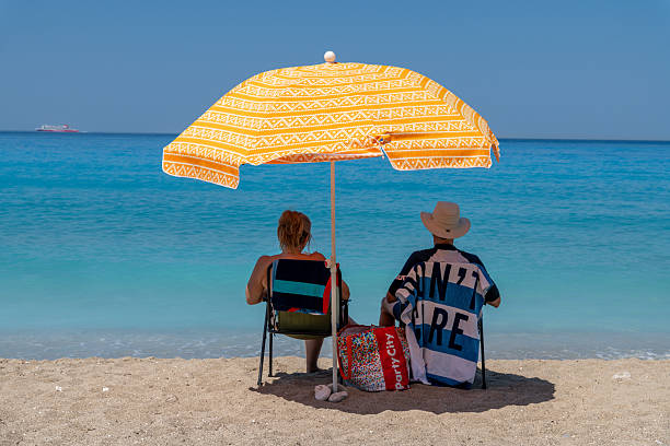 Nydri - Ionian Islands - Greece - 08-09-2025: Two people sitting under a patterned orange umbrella on sandy beach, facing calm turquoise sea with ship in distance under clear sky.