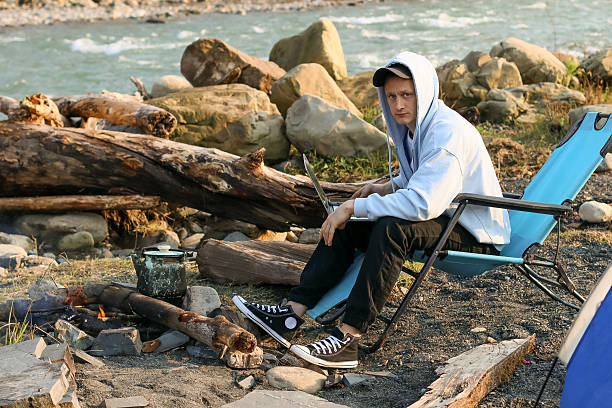 A young man in a light hoodie lounges in a folding chair with a laptop on his lap, beside a small campfire and blue tent on a rocky riverbank.