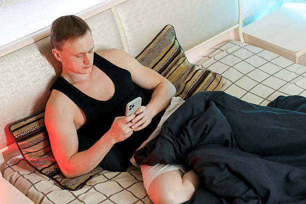 Man resting on bed with hands over stomach, enveloped in blanket under warm bedroom lighting.