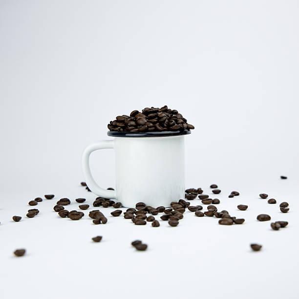This studio-shot image presents a clean and minimalist arrangement of dark roasted coffee beans surrounding a white ceramic mug that is filled to the brim. The scattered beans create a natural composition that highlights texture, aroma, and the handcrafted nature of coffee culture. The neutral background enhances the contrast between the glossy beans and the matte mug, making the scene suitable for concepts related to roasting, specialty coffee, café branding, product advertising, or packaging layouts. The simple setup also evokes themes of morning rituals, artisanal brewing, and modern beverage aesthetics.