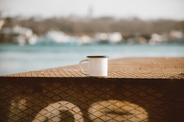 This image features a white enamel coffee mug placed on a weathered wooden deck with a view of a large bridge and the waterfront in Istanbul. The muted tones and soft natural light emphasize a calm outdoor atmosphere, while the distant city structures highlight the blend of modern architecture and scenic views that characterize the area. The composition conveys a peaceful moment by the water, making it suitable for visual themes such as Istanbul travel culture, slow living, urban relaxation, and serene morning routines.