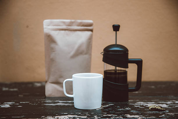 This image displays a simple arrangement of a plain kraft paper coffee bag, a white ceramic mug, and a black French press placed on a weathered wooden table. The warm background and rustic textures create an inviting café-like atmosphere focused on slow brewing and minimalist design. The combination of packaging, drinkware, and brewing equipment reflects themes of handcrafted coffee culture, artisanal preparation, and modern branding aesthetics. This setup is suitable for showcasing product packaging, illustrating brewing concepts, or supporting lifestyle content related to specialty coffee and contemporary cafe environments.