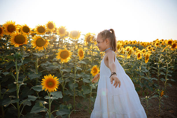 Beautiful little girl in yellow garden of sunflowers. A fair-haired 7-year-old girl in a yellow linen dress stands in a field of sunflowers, in the photo there is a summer sunset in the field