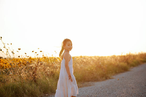 A pretty little girl is picking wildflowers in a lush meadow at sunset. She's alone in a peaceful natural setting, with a forest in the background.