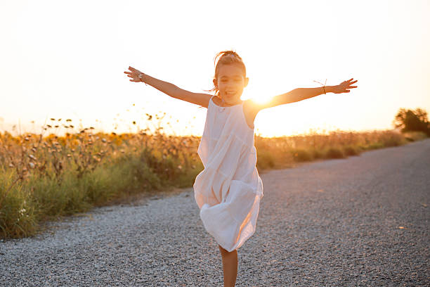 A pretty little girl is picking wildflowers in a lush meadow at sunset. She's alone in a peaceful natural setting, with a forest in the background.