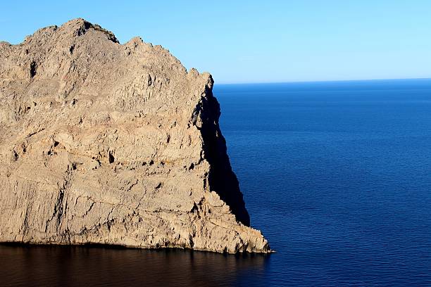 Minimalist view of a towering limestone cliff at Cap Formentor, Mallorca, dropping into deep blue Mediterranean waters under clear sky, showcasing dramatic geology and pristine coastal scenery.