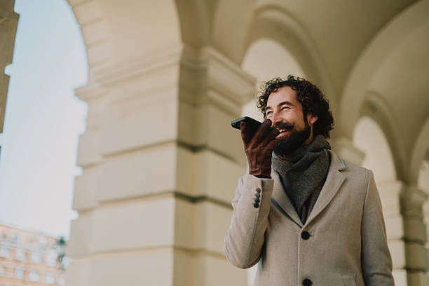A stylish, adult man with a beard wears a beige coat and scarf, smiling as he holds a phone under a graceful archway in a winter urban setting, conveying confidence, warmth, and holiday mood.