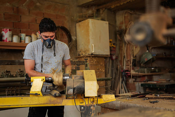 Carpenter wearing a protective mask works diligently with a lathe in a cluttered workshop, with wood shavings swirling around, showcasing the art of traditional woodworking