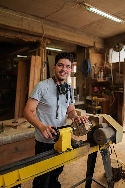istock Young carpenter smiling while turning a piece of wood on a lathe in a dusty, old workshop, surrounded by sawdust and tools, showcasing craftsmanship and creativity in woodworking 2247696209
