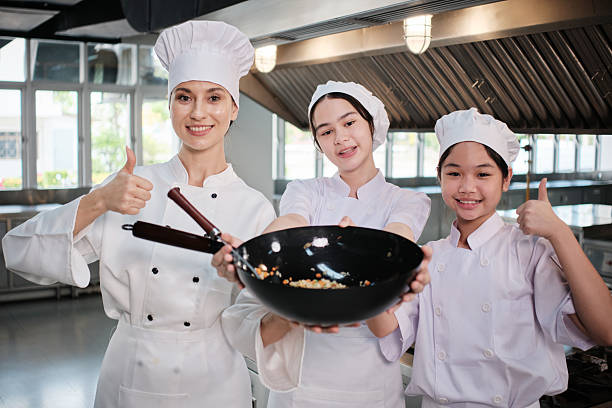 Teenage trainee girls in white aprons are proud to show fried rice in pan, cooking class work with master chef in gourmet restaurant kitchen, delicious cuisine with ingredients, and culinary meals.