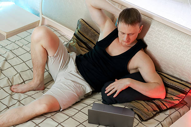 istock Arm raised man reclining on pillows with laptop on bed in ambient glow 2247732483