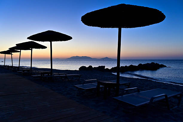 Two deck chairs on empty beach under straw umbrella in front of blue ocean.