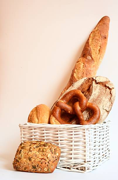 Assorted artisan bakery bread arranged in a white basket with a clean background. Includes rustic loaf, pretzel, baguette, and seed roll. Great for culinary and food industry use.