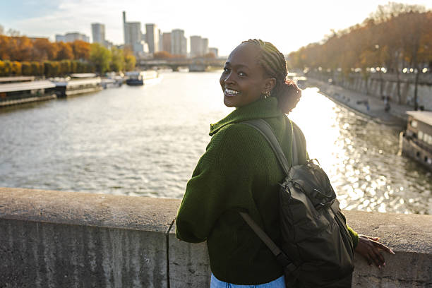 A cheerful woman with a green sweater and backpack gazes over the Seine in Paris, enjoying a sunny day and city views along the riverfront.