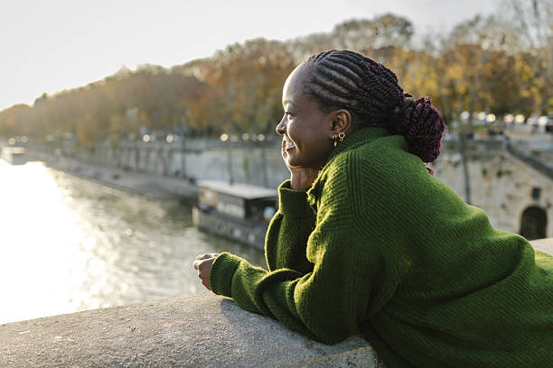 A stylish woman with braided hair wearing a green sweater relaxes on a Paris riverside, basking in sunlight.