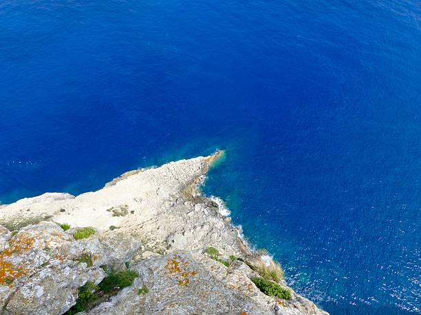 A dramatic view from high above the coastline of Mallorca, showcasing rugged cliffs covered in lush green vegetation. Below lies the calm and expansive Mediterranean Sea, stretching toward the horizon. The peaceful atmosphere, natural textures, and vibrant colors highlight the untouched beauty of this island landscape.