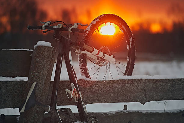 A bicycle is hung on a wooden fence in the village against the backdrop of a bright sunset sky in winter and snow