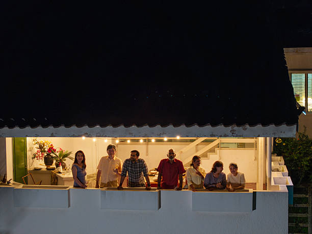 Asian  group of people enjoying a relaxed evening gathering on a home balcony, standing together and chatting under warm lights, viewed from outside at night. Kuala Lumpur.