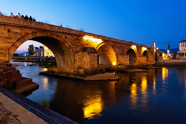 Historic Stone Bridge in Skopje with glowing street lamps reflecting in Vardar River on summer evening, surrounded by illuminated architecture