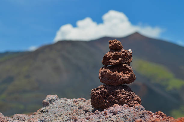 Balanced volcanic lava rocks forming cairn in foreground with Mount Etna volcano and white clouds in background. Natural meditation and travel concept in Sicily, Italy.