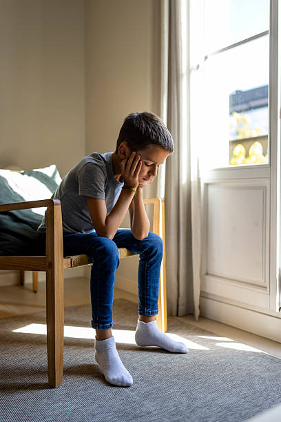 istock Young Boy Sitting Alone on Chair by Window, Looking Down, Feeling Sad at Home 2247762279