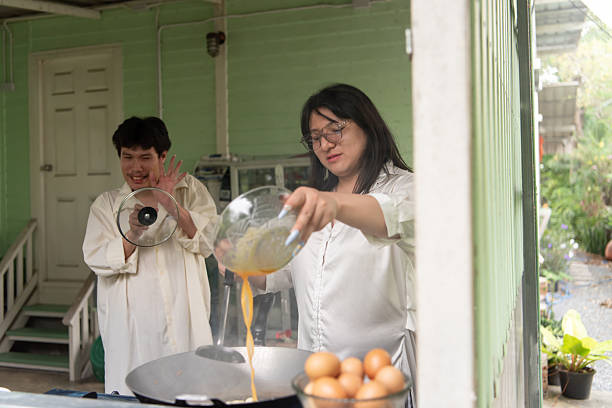LGBTQ friends, one with long hair and one with short, share a joyful moment preparing a Thai-style omelette for breakfast in a cozy open kitchen. The scene captures the fun of cooking together.