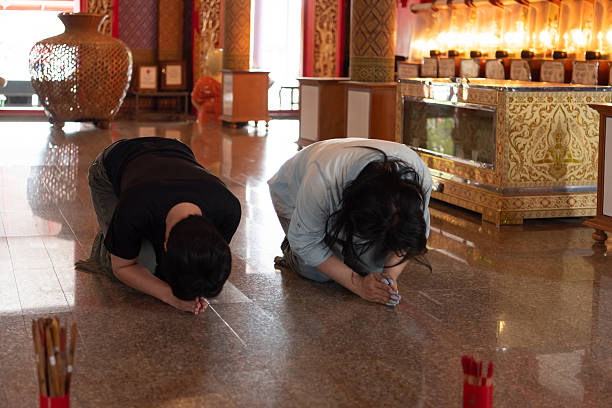 Two LGBTQ friends are kneeling and praying at a Thai temple as part of a religious merit-making ritual, offering incense and flowers as they engage in Buddhist practices.