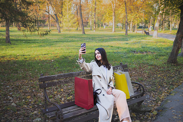 istock Beautiful young woman taking selfies during a walk on a beautiful autumn day using mobile phone in the park full of fallen autumn leaves after shopping carrying shopping bags. 2247767007