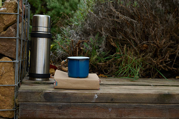 A simple rustic outdoor setup features a metal thermos, a blue enamel mug, and a closed book on a wooden deck beside greenery.