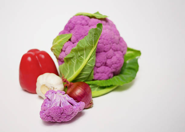 Colorful assortment of fresh vegetables including purple cauliflower, red bell pepper, garlic and red onion on a white background. Composition for healthy food themes.
