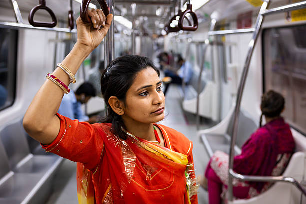A young woman in a vibrant red-orange traditional outfit stands inside a modern metro car, holding an overhead strap. She looks thoughtfully ahead, her bangles and small earrings visible, while other passengers sit blurred in the background. The clean stainless handrails, hanging grips and empty seats frame this quiet, candid moment during a commute.