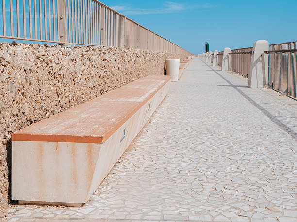 A long stone bench stretches along a peaceful coastal pathway, with bright blue skies and calm ocean waves in the background. Ideal spot for relaxing and enjoying the view.