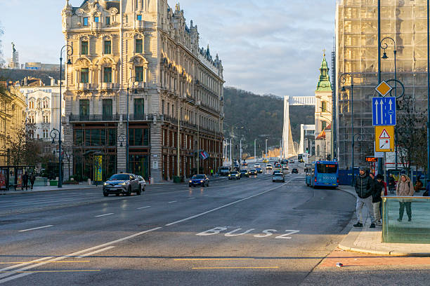 Morning traffic on Szabad Sajtó Blvd toward Elisabeth Bridge, historic Ferenciek Square buildings lit by winter sun, central Budapest commute.