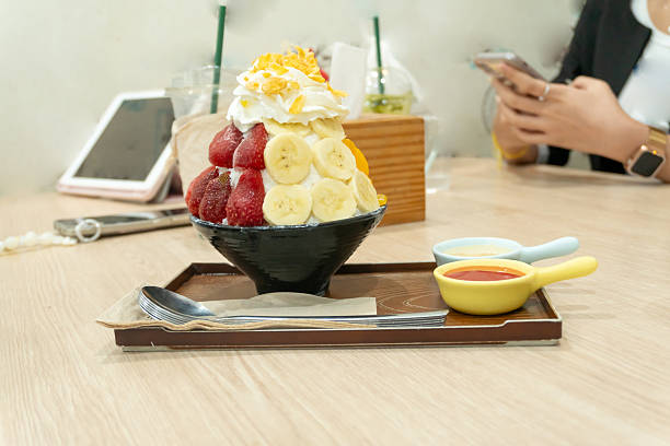 A set of Korean Bingsu in small black bowl with several fruits topping and red sauce with sweeten condensed milk in mini plastic bowls in brown wooden tray served customer in cafe or restaurant