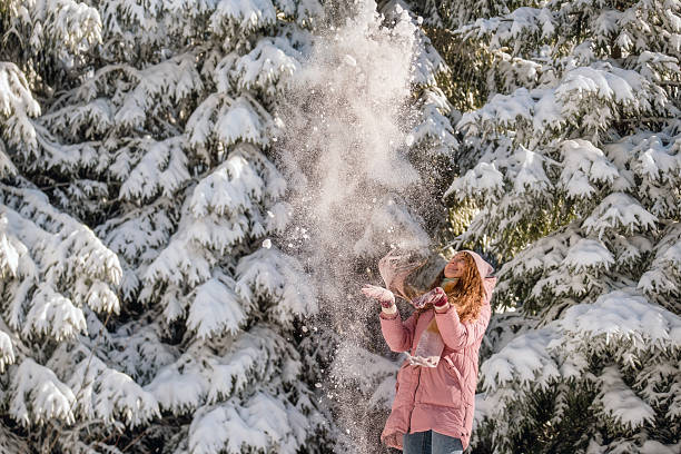 Young women having fun in snow winter forest. Female in cozy scarf and pink jacket throws snow up, enjoying first snow among snowcapped fir trees. Happy wintertime and Christmas holidays.