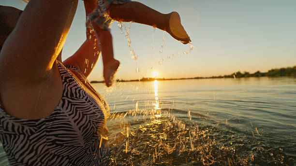 Caucasian mother joyfully lifting her toddler in the lake during sunset, creating a playful and warm moment with water splashing around them.