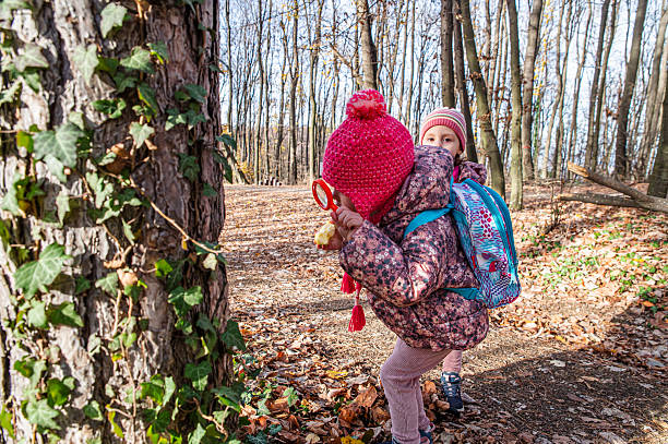 Two kids wearing colorful winter outfits are exploring nature in a serene forest, showcasing curiosity and adventure amidst autumn leaves and trees.