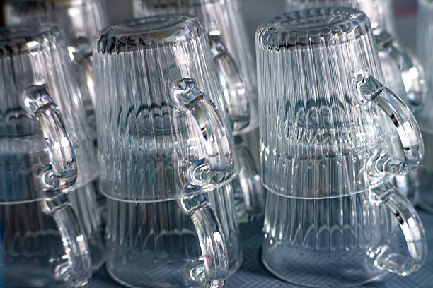 Transparent glass mugs stacked upside down on a tray in a neat arrangement. Close-up shot highlighting light reflections and textures of clear glass.