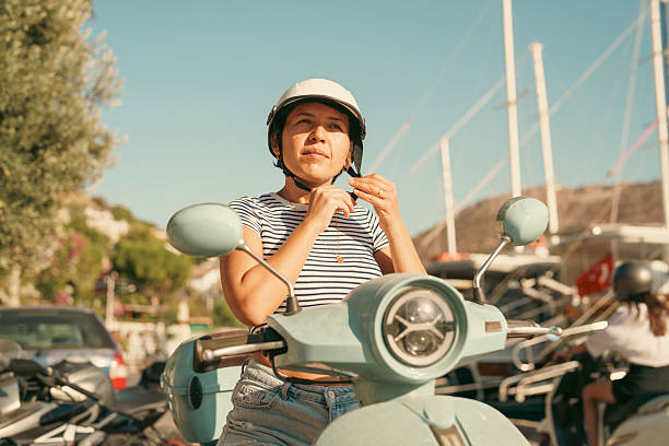 Front view of a woman on a parked scooter adjusting her helmet strap. The scene is outdoors with boats, vehicles, and marina elements visible in the background.