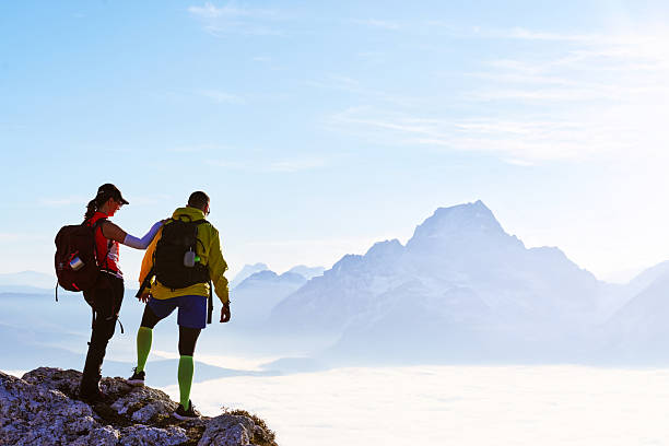 Two hikers stand on a rocky summit, gazing at the stunning landscape below. The sun sets in the distance, casting a warm glow on the clouds that blanket the valley.Couple enjoying breathtaking view from mountain peak during sunset on a clear day