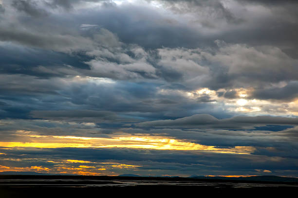 Under a vast expanse, clouds swirl above Iceland as the sun sets, casting golden hues against the turbulent sky. The landscape below reflects the shifting light, evoking a sense of wonder.