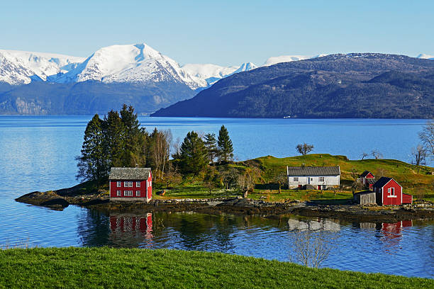 Picturesque spring scenery in Hardanger, in Western Norway, with calm sea, blue sky, snowcovered mountains in background and a charming islet in the middle