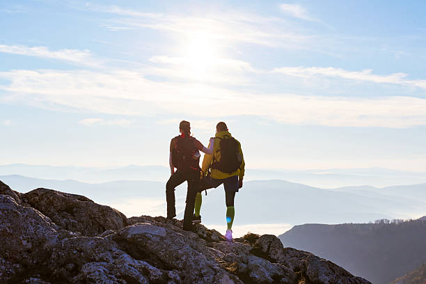 Two hikers stand on a rocky summit, gazing at the stunning landscape below. The sun sets in the distance, casting a warm glow on the clouds that blanket the valley.Couple enjoying breathtaking view from mountain peak during sunset on a clear day