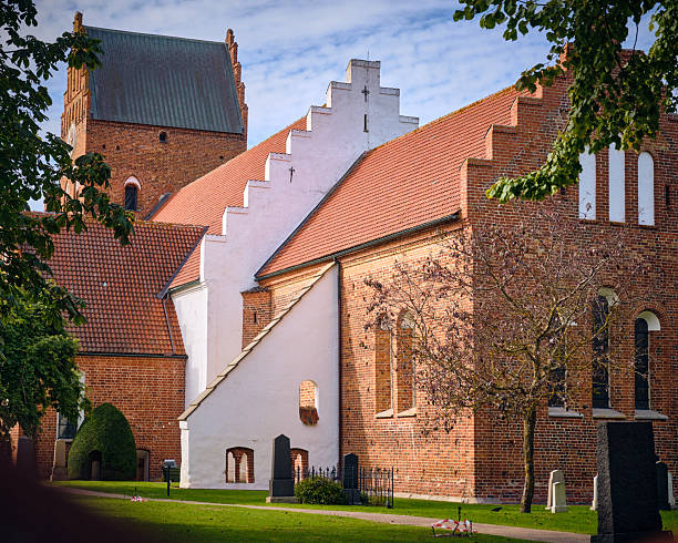 Ahus, Sweden; September 22, 2025: Brick Gothic church exterior with stepped gables and red tile roofs framed by trees and graveyard elements under soft daylight.