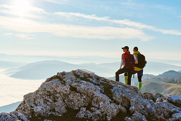 Two hikers stand on a rocky summit, gazing at the stunning landscape below. The sun sets in the distance, casting a warm glow on the clouds that blanket the valley.Couple enjoying breathtaking view from mountain peak during sunset on a clear day